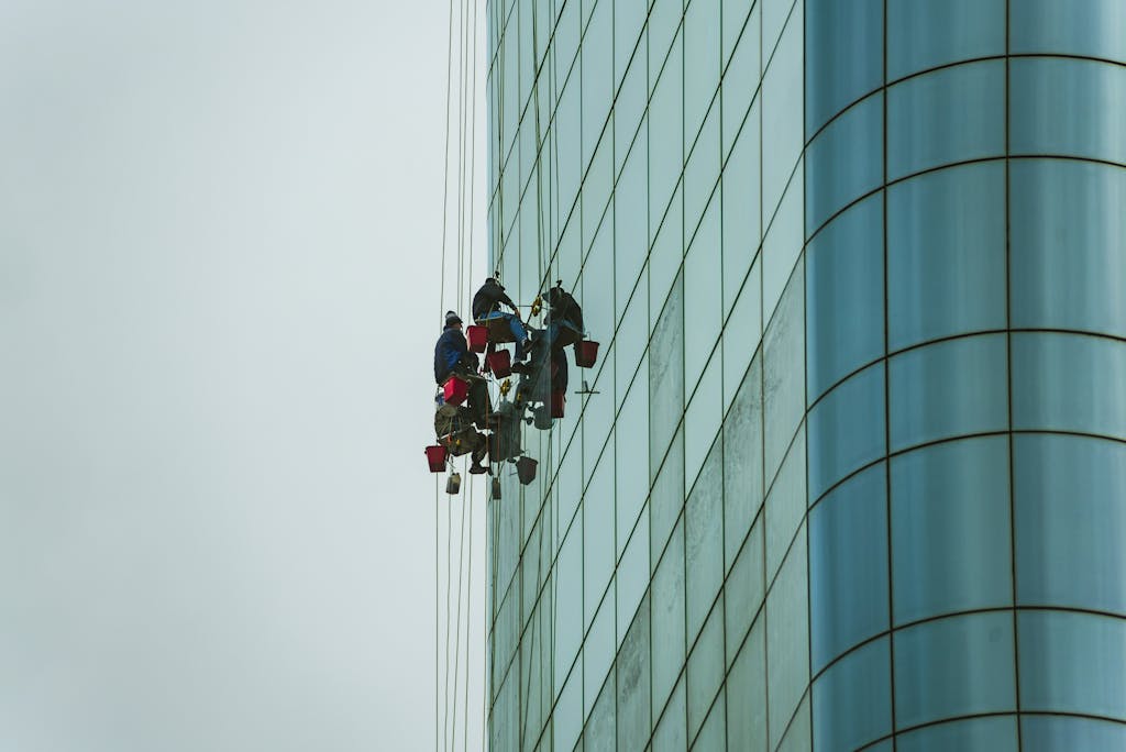 Window cleaners suspended on ropes working on a tall glass building facade.